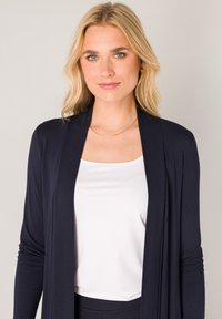 Marineblauwe cardigan gemaakt van zachte stof, met een open voorkant, valt tot halverwege de dij, gecombineerd met een witte nauwsluitende tanktop. Goudkleurige ketting als accent.