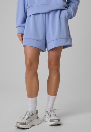 Osoba ubrana w jasnoniebieskie szorty i pasującą bluzę, białe sportowe skarpetki oraz białoszare buty do biegania stoi na szarym tle.