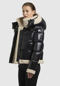 Zwarte pufferjack met een glanzende afwerking, beige faux bont voering, oversized kraag en drukknoopsluitingen bij de hals en manchetten.