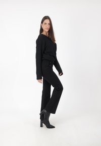 Maglione nero lavorato a maglia con scollatura ampia, abbinato a pantaloni neri a vita alta e stivaletti neri con tacco, su uno sfondo bianco semplice.