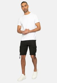 Weißes T-Shirt mit Rundhalsausschnitt kombiniert mit schwarzen Cargo-Shorts, die über Seitentaschen und Kordelzüge am Saum verfügen. Getragen mit weißen Sneakers.