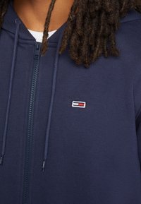 Felpa con zip blu navy realizzata in tessuto morbido, con cerniera frontale, lacci e un piccolo logo ricamato in rosso e bianco.