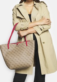 Bežová taška přes rameno s texturovaným vzorem a červenými syntetickými koženými popruhy. Na přední straně se nacházejí stříbrné detaily a logo.