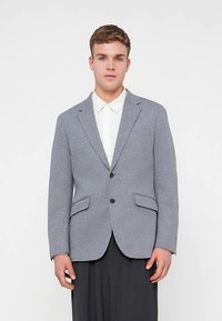 Grijze geweven blazer met twee buitentassen, enkele knoopsluiting en steekkraag, gecombineerd met een wit overhemd met kraag eronder.