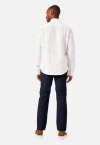 Man die wegkijkt, gekleed in een wit longsleeve shirt, donkere marineblauwe broek en bruine suède schoenen met witte zolen tegen een effen achtergrond.