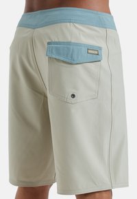 Svetlo béžové plavky s modrým pásom. Obsahujú zadnú kapsu s chlopňou, viditeľné švy a hladkú textúru.