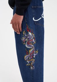 Denimjeans med en flerfärgad broderad drake-design på vänster lår, med flammande detaljer i rött och gult, samt en bakfickdetalj.