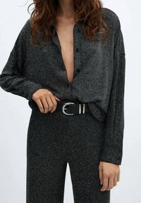 Femme portant une chemise à boutons noire pailletée avec un décolleté plongeant, rentrée dans un pantalon assorti, tenant une ceinture noire avec une boucle argentée.