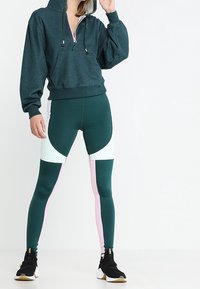 Felpa con cappuccio in tessuto verde scuro con una cerniera rosa. Leggings alto vita con un design a blocchi di colore in verde scuro, bianco e rosa.