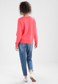 Felpa corallo con scollatura rotonda, maniche lunghe e dettagli discreti sul retro. Abbinata a jeans azzurri e sneakers bianche.