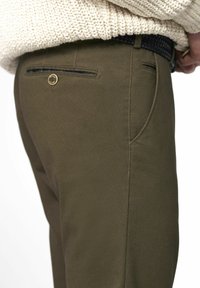 Pantalons vert olive avec un tissu texturé, comportant une poche arrière ornée d'un bouton en or décoratif et une ceinture tissée partiellement visible.