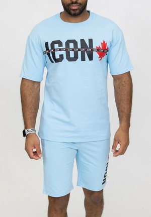 Homme portant un ensemble short et t-shirt bleu clair avec le texte "ICON" et une feuille d'érable rouge, debout devant un fond blanc uni.