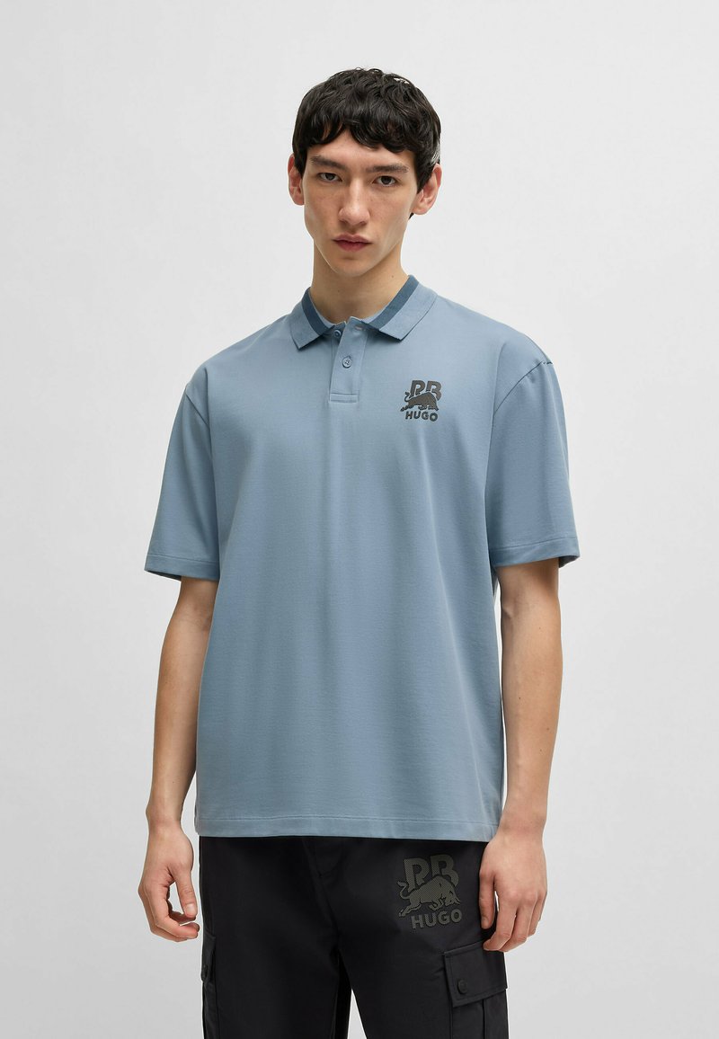 Paita polo dalam kain biru muda, dilengkapi dengan kerah, placket dua tombol, dan logo hitam di dada. Lengan pendek dengan potongan yang nyaman.