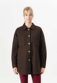 Giovane donna che indossa un cappotto button-up oversized marrone scuro con grandi tasche frontali e pantaloni rossi, in piedi su uno sfondo bianco uniforme.