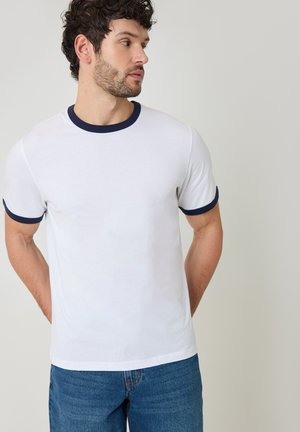 Uomo con capelli ricci e scuri, indossa una maglietta bianca con colletto e bordi delle maniche blu navy, jeans blu, in piedi con le mani dietro la schiena.
