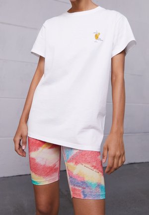 T-shirt bianco in cotone con maniche corte, con una grafica di un cocktail giallo. Abbinato a pantaloni bici colorati con un motivo astratto accompagnato da del testo.