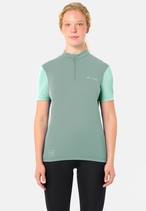 Vrouw met krullend haar die een kortemouwen wielershirt draagt in twee tinten groen en zwarte legging, staand tegen een witte achtergrond.