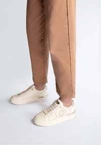 Beige sneakers med et struktureret mønster, snørebåndsdesign og metalliske accenter på hælen. Parret med lysebrune, tætsiddende bukser.