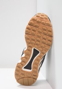 Černobílá podrážka atletické boty s gumovým trakčním vzorem, světle hnědými vložkami a logem Torsion uprostřed. Texturovaný povrch.