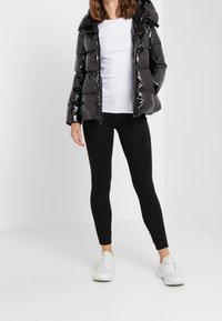 Kobieta w błyszczącej czarnej kurtce puchowej, białej koszulce, czarnych legginsach i białych trampkach stojąca na jednolitym białym tle.