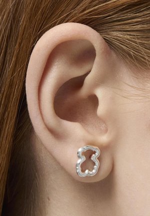 Pendiente de plata en forma de oso, con un diseño calado. Llevado en el lóbulo de la oreja, mostrando la superficie lisa y reflectante del pendiente.