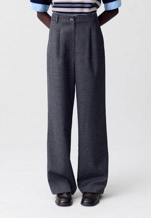 Pantalons larges gris en tissu texturé, avec une fermeture à bouton à l'avant et des plis, associés à un haut rayé et des chaussures noires.