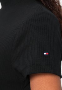 Gros plan d'un t-shirt noir côtelé à manches courtes porté par une personne, avec un petit logo rectangulaire rouge, blanc et marine sur la manche.