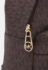 Zaino marrone a fantasia con materiale testurizzato, un tirazip dorato e un logo circolare MK ben visibile.
