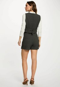 Donna di spalle che indossa un gilet senza maniche grigio scuro e pantaloni corti abbinati, con una camicia a maniche lunghe bianca e sandali alti marroni.