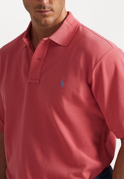 Κόκκινο πόλο μπλουζάκι από υφασμάτινο ύφασμα, με κλασικό γιακά, τριπλό κουμπί και μικρό μπλε κεντημένο λογότυπο στο στήθος.