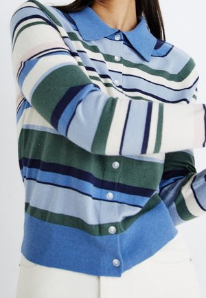Femme portant un cardigan rayé boutonné avec des rayures horizontales bleues, vertes, blanches et bleu marine et un col bleu.