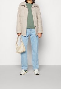 Veste beige matelassée, sweat-shirt vert, jean bleu clair et baskets blanches avec des accents verts. Tenant un sac à main beige.