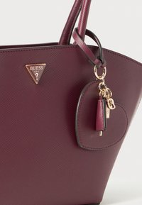 Burgunderfarget teksturert håndveske med doble håndtak, gullfarget GUESS-logo i trekantform, og hjerteformet charm med nøkkel og metallmerker festet.