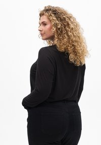 Svart långärmad topp i mjuk, elastisk tyg, kombinerad med svarta byxor. Lockigt hår ramar in ansiktet och visar en avslappnad passform.