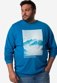 Blaues Langarmshirt mit einem Bergmotiv in hellen Farben und dem Text "DIE BERGE RUFEN ZU GIPFELABENTEUERN" am unteren Rand.