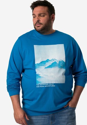 Blaues Langarmshirt mit einem Bergmotiv in hellen Farben und dem Text "DIE BERGE RUFEN ZU GIPFELABENTEUERN" am unteren Rand.