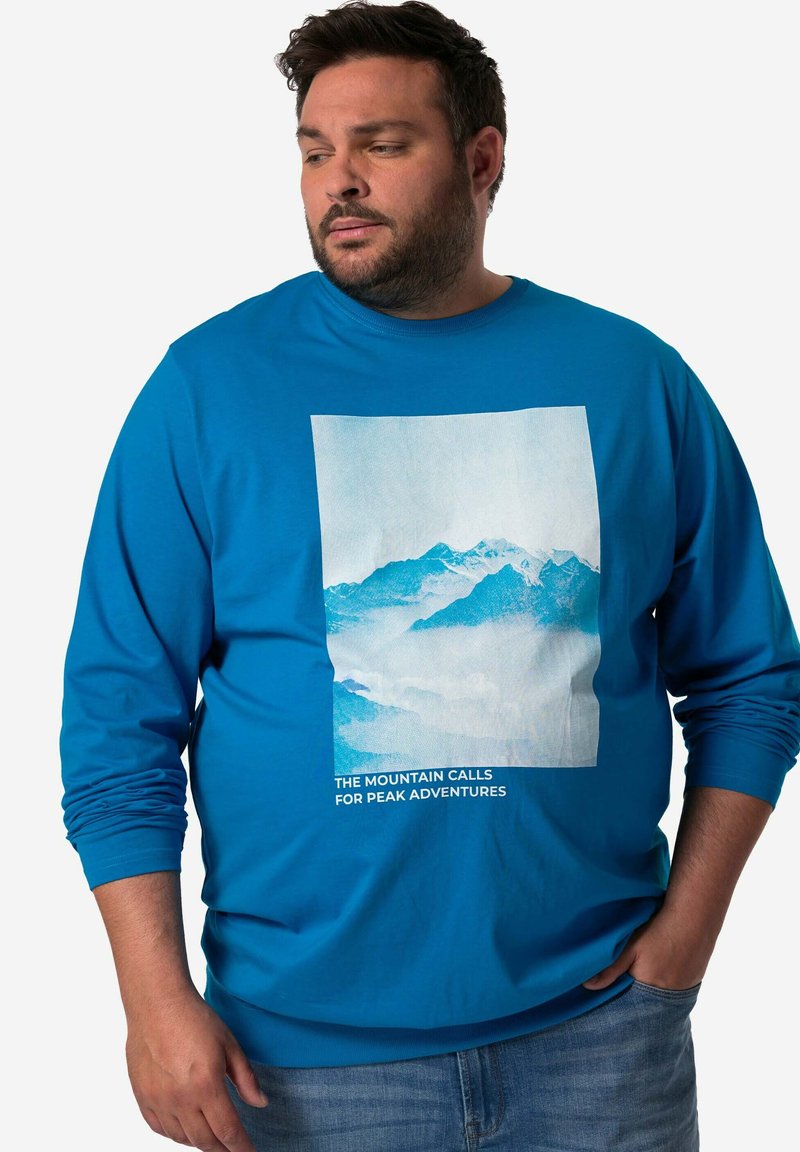 Blaues Langarmshirt mit einem Bergmotiv in hellen Farben und dem Text "DIE BERGE RUFEN ZU GIPFELABENTEUERN" am unteren Rand.