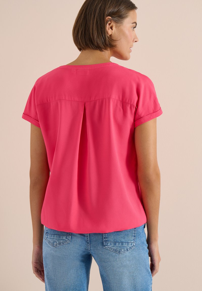 Femme aux cheveux courts bruns portant un chemisier rose vif à manches courtes et un jean bleu, vue de dos sur un fond beige uni.