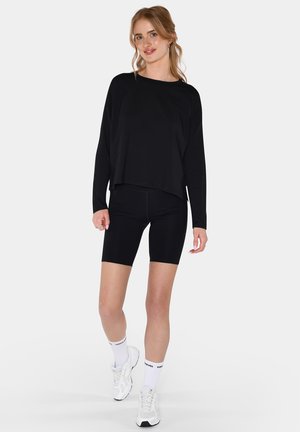 Kvinne med lyst hår iført en svart langermet topp, svarte sykkelshorts, hvite sokker og hvite joggesko, stående på hvit bakgrunn.