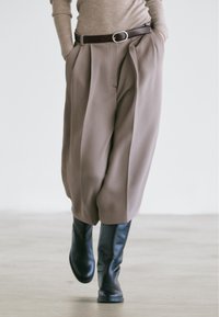 Beige pleated trousers with side pockets, cinched at the waist by a brown leather belt, paired with knee-high black leather boots.