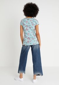 Camiseta floral de color azul claro con mangas cortas, que presenta patrones de flores en rosa y verde. Combinada con jeans de denim desgastados de pierna ancha y zapatillas blancas.