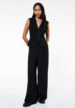 Donna in piedi con le mani dietro la schiena che indossa un gilet nero senza maniche con bottoni, pantaloni neri a gamba larga e scarpe nere su sfondo bianco.