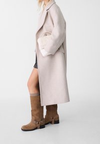 Persona che indossa un lungo cappotto beige, stivali marrone scamosciato fino al ginocchio, e tiene in mano una piccola borsa di pelliccia bianca su uno sfondo neutro.
