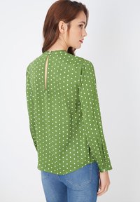 Blusa verde con lunares blancos, escote redondo, mangas largas y detalle de abertura en la espalda. Tela texturizada que cae suavemente.