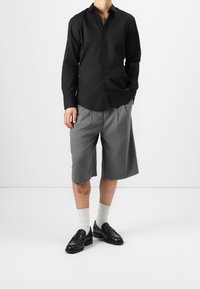Schwarzes Langarmshirt, graue gefaltete Shorts, weiße Socken, schwarze Lederschuhe. Lässiges Outfit mit kontrastierenden Texturen und Farben.