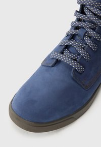 Shoe azul de camurça com atacadores azuis e bege estampados e sola de borracha castanha, sobre um fundo branco.