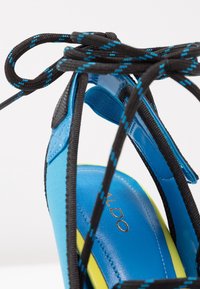 Sandale bleue et jaune avec des lacets noirs et bleus noués en nœud au-dessus de la bride de cheville, montrant une partie de la semelle intérieure avec un texte doré.