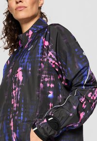 Veste légère noire avec un motif abstrait vibrante en rose et bleu, fermeture éclair, col haut et poignets élastiques. Comprend des accents réfléchissants.