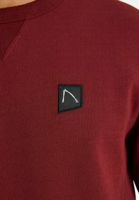 Felpa bordeaux con colletto a girocollo a coste, tessuto testurizzato e una toppa quadrata nera con un design geometrico bianco sul petto.