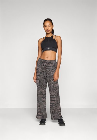 Sujeción deportiva negra con un logo, combinada con pantalones sueltos y estampados que presentan una textura de cocodrilo en tonos de gris y púrpura.